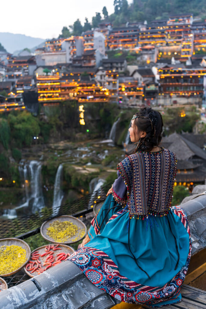 Joven china sentada mirando paisaje iluminado contenta con nuestra logística de viajes y vestida con ropa tradicional de Lijiaang